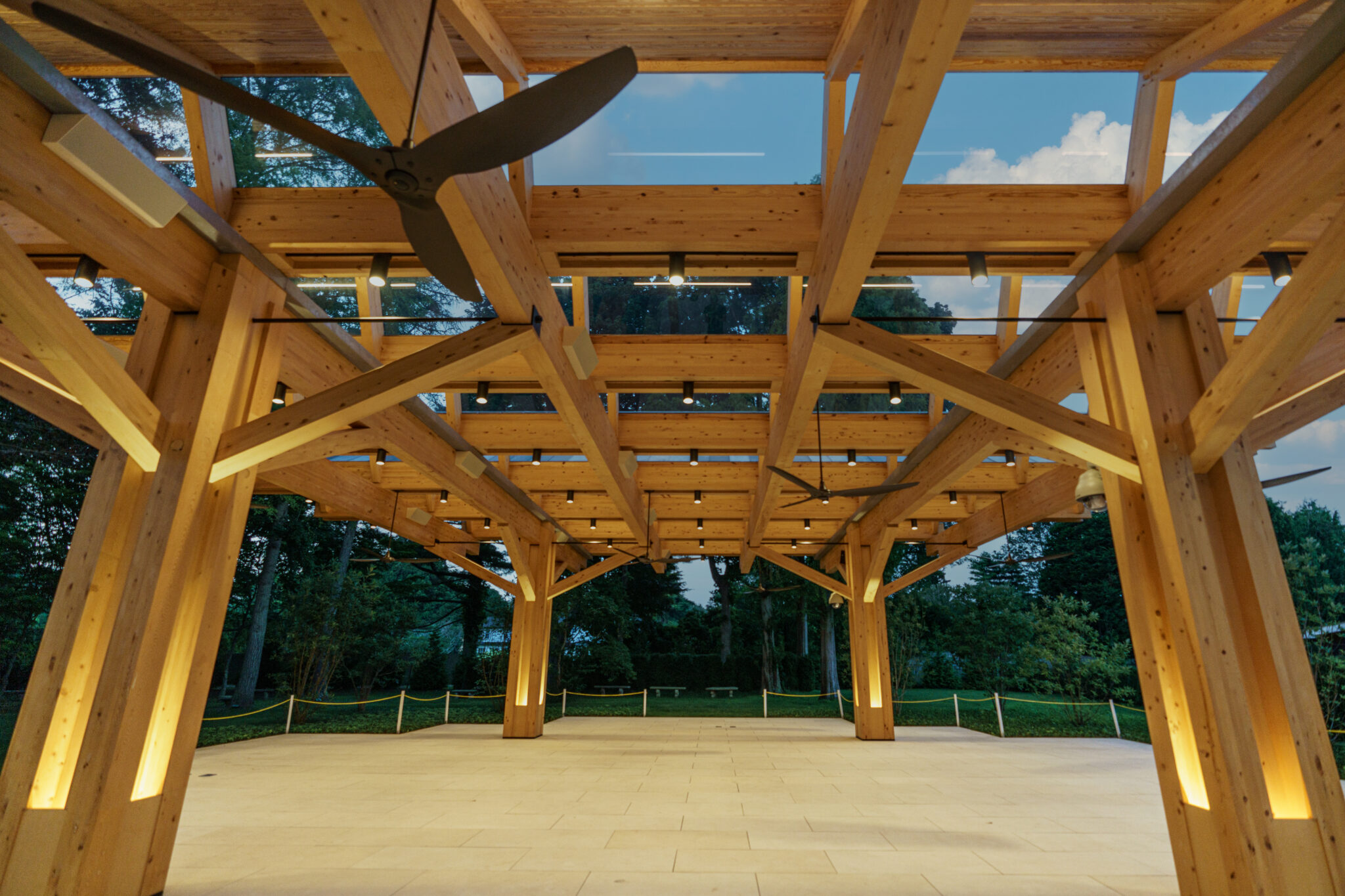 An open-air timber frame pavilion with a grid of ceiling lights, ceiling fans, and a light-colored paved floor, overlooking trees and grass.