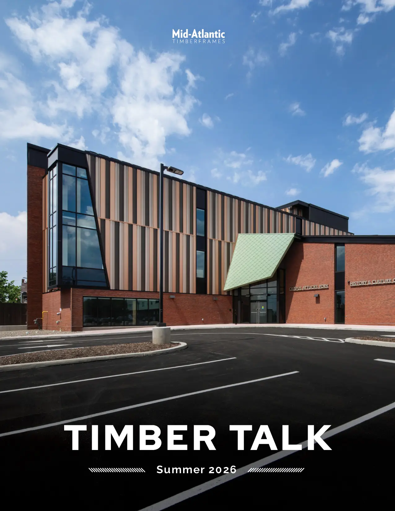 A modern building with brick and vertical wood panels sits under a blue sky. The cover text reads TIMBER TALK Summer 2026 and Mid-Atlantic Timberframes at the top. The empty parking lot is in front.