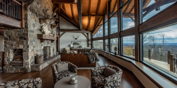 Rustic lodge living room with a stone fireplace, vaulted timber ceilings, and a wall of windows overlooking a forest valley.